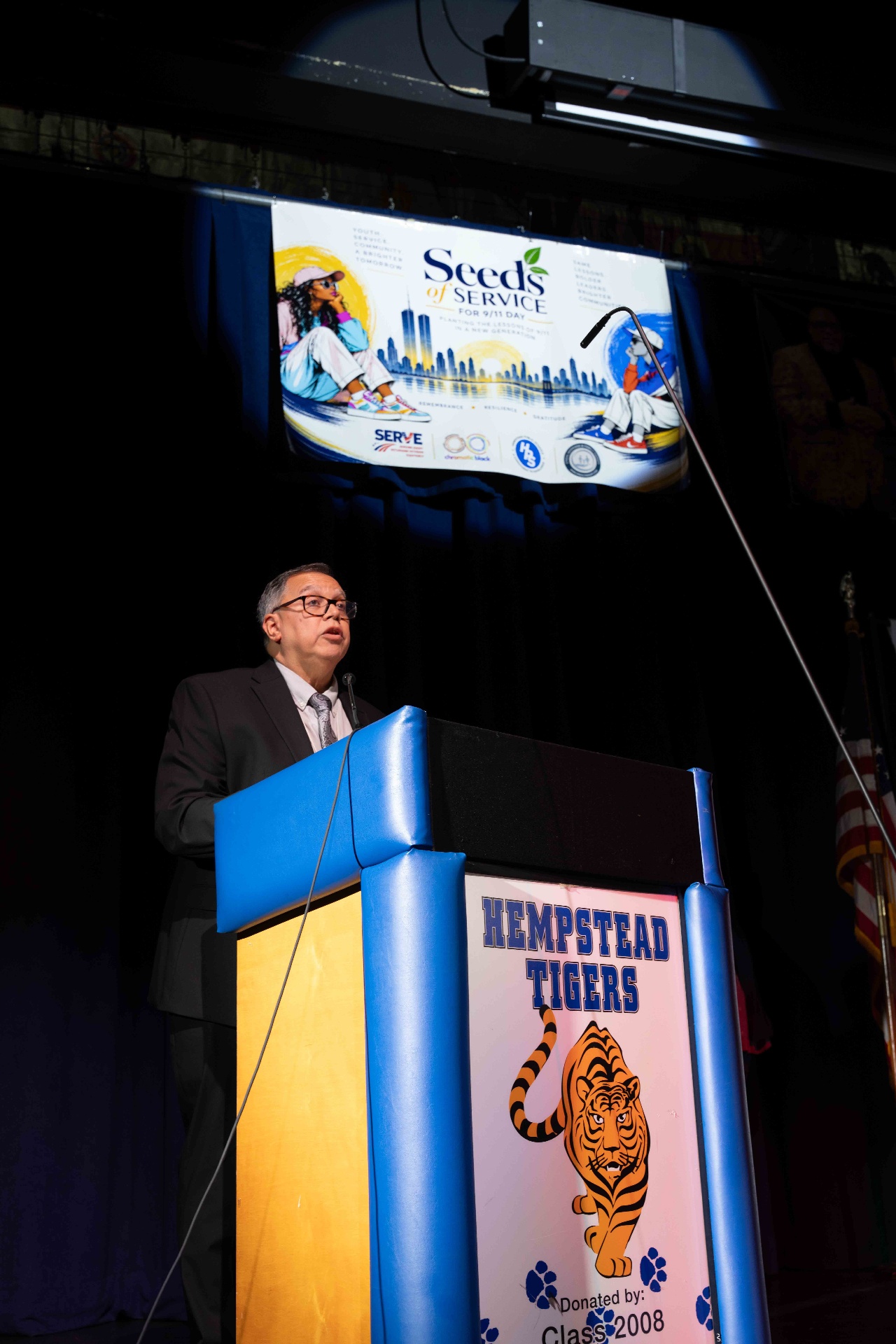 A speaker addressing students from the podium beneath the Seeds of Service banner