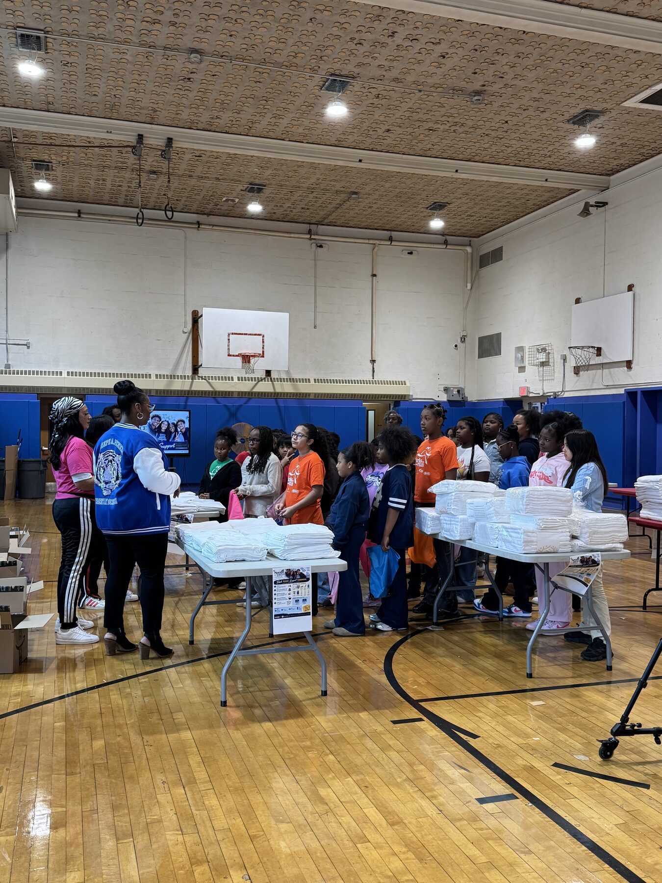 Students standing behind long tables of supplies as an adult gives instructions in the Joseph A. McNeil Elementary School gymnasium